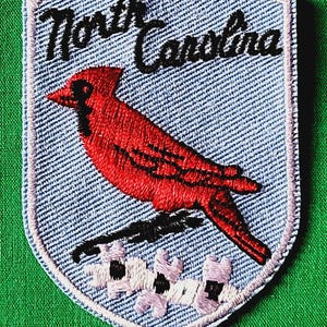 May include: A shield-shaped embroidered patch with a light blue background featuring a red cardinal bird perched on a branch above white dogwood flowers. The text North Carolina is stitched in black script across the top of the patch.