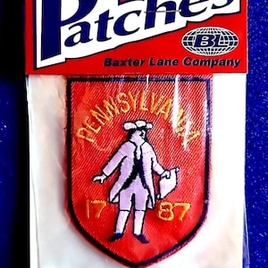 May include: Iron-on patch with a red shield design featuring a colonial figure and the text "PENNSYLVANIA 1787". The patch is packaged in a clear plastic bag with a red and white header that reads "Patches" and "Baxter Lane Company".