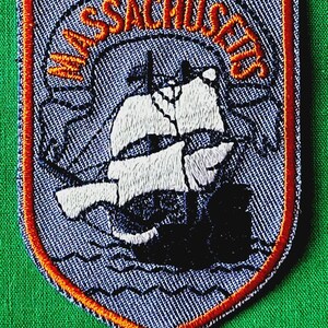 May include: A shield-shaped embroidered patch featuring a light blue background with a dark orange border. The patch displays a white and black sailing ship graphic and the text MASSACHUSETTS arched in orange lettering across the top section.