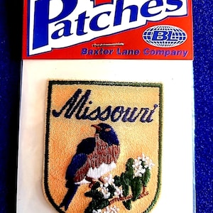 May include: Iron-on patch featuring a shield design with the word "Missouri" in navy blue script. The patch showcases a bluebird perched on a flowering branch, with a gold background and green border. The packaging reads "Patches" and "Baxter Lane Company."