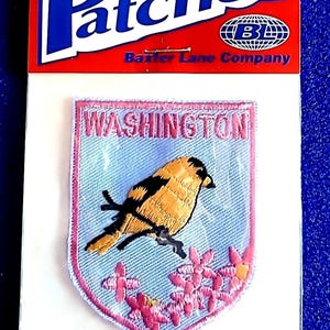 May include: Embroidered patch featuring a yellow and black bird perched on a branch with pink flowers, set against a light blue background. The patch is labeled "WASHINGTON" and is framed with a pink border. The top of the packaging reads "Patches".