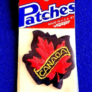 May include: An iron-on patch featuring a red maple leaf with the word "CANADA" in yellow lettering. The patch is outlined in black and is on a red and white backing. The packaging reads "Patches" and "Made in U.S.A. by Voyager."
