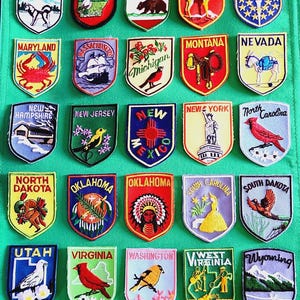 May include: A collection of twenty-five embroidered state patches arranged in five rows on a green fabric background. Each shield-shaped patch features a unique illustration representing a U.S. state, including Alaska, Arkansas, California, Colorado, Indiana, Maryland, Massachusetts, Michigan, Montana, Nevada, New Hampshire, New Jersey, New Mexico, New York, North Carolina, North Dakota, Oklahoma, South Carolina, South Dakota, Utah, Virginia, Washington, West Virginia, and Wyoming.