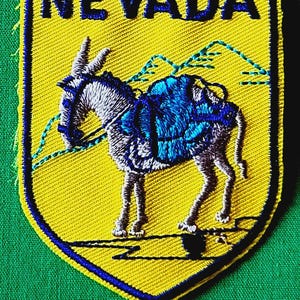May include: A shield-shaped embroidered patch with a yellow background and a dark blue border. It features a grey donkey carrying a blue pack, with blue mountain outlines in the background and the word NEVADA stitched in dark blue capital letters.
