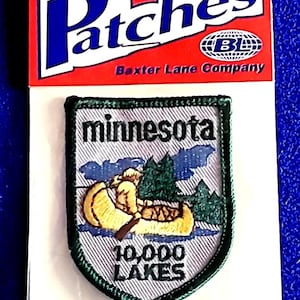 May include: Iron-on patch featuring the word "Patches" in white and blue on a red background. The patch has a Minnesota theme, with a canoe, trees, and the text "10,000 Lakes". The patch is bordered with green stitching.