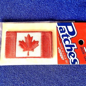 May include: An iron-on patch featuring the Canadian flag, with a red maple leaf on a white background, framed by red borders. The patch is packaged with a red and blue label that reads "Patches" and "Iron-On".
