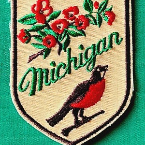 May include: A shield-shaped embroidered patch with a tan background and a black border. It features a red apple blossom branch with green leaves, the word Michigan in green cursive script, and a black and red robin bird perched on a branch.