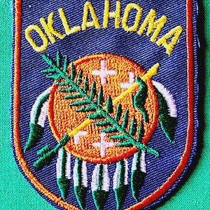 May include: A shield-shaped embroidered patch featuring the word OKLAHOMA in yellow thread at the top. The center displays an orange circular design with a green olive branch and a crossed peace pipe and arrow, bordered by red and blue edges.