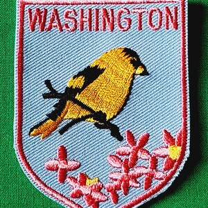 May include: A shield-shaped embroidered patch with a light blue background and a red border. It features the word WASHINGTON in red capital letters at the top, a yellow and black goldfinch perched on a branch, and pink flowers at the bottom.