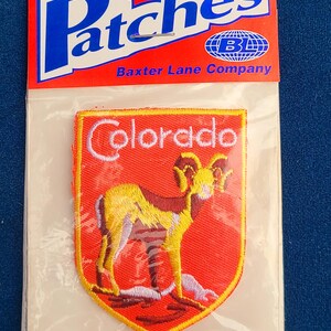 May include: An iron-on patch featuring a golden ram on a red background with the word "Colorado" above. The patch is in a clear plastic package with a red and white header that reads "Patches" and "Iron-On".