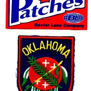 May include: An iron-on patch featuring the word "Oklahoma" in yellow, with a shield design. The patch includes a red circle with a cross and a green sprig, and is bordered in blue and red.