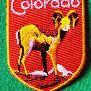 May include: A shield-shaped embroidered patch with a red background and a yellow border. It features a bighorn sheep standing on a rocky base with white snow accents. The word Colorado is stitched in white cursive lettering at the top of the patch.