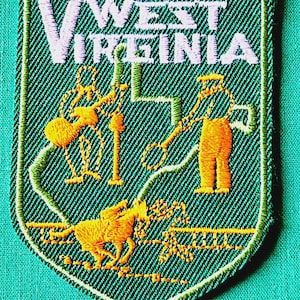 May include: A shield-shaped embroidered patch in dark green with a light green border. It features the white text "WEST VIRGINIA" at the top, with orange embroidered figures depicting a person playing a guitar, a person with a hoop, and a person on a horse.