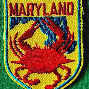May include: A shield-shaped embroidered patch featuring a bright yellow background with a blue border. The word MARYLAND is stitched in red capital letters at the top. A large red crab is centered on the patch over a blue graphic design element.