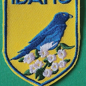 May include: A shield-shaped embroidered patch with a yellow background and a blue border. The text IDAHO is stitched in blue at the top. A blue mountain bluebird is perched on a cluster of white flowers with green leaves in the center.