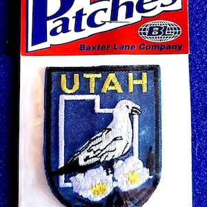 May include: Iron-on patch featuring the word "Patches" in white and blue on a red background. The patch itself is a blue shield shape with the word "UTAH" in gold, a white bird, and white and yellow flowers.