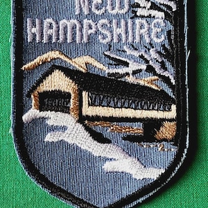 May include: A shield-shaped embroidered patch featuring a winter scene with a covered bridge, snow-covered ground, and bare trees. The text NEW HAMPSHIRE is stitched in light blue at the top against a dark blue background with a black border.