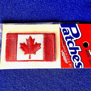 May include: A rectangular embroidered patch featuring the Canadian flag with a red maple leaf on a white background flanked by two red vertical bars. The patch is attached to a red and white cardboard backing labeled Patches, Iron-On, Voyager.
