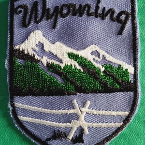 May include: A shield-shaped embroidered patch with a light blue background. The text Wyoming is stitched in black at the top. Below, white snow-capped mountains sit above green pine trees. A white wooden fence is featured at the bottom of the patch.