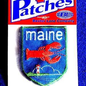May include: An iron-on patch featuring a blue shield with a red lobster and the word "maine" in white. The patch is on a red and white backing that reads "Patches" and "Iron-On".