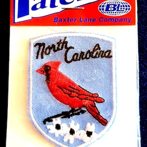 May include: Iron-on patch featuring a red cardinal bird and the words "North Carolina" embroidered on a light blue shield. The patch is packaged on a white card with the word "Patches" in blue.