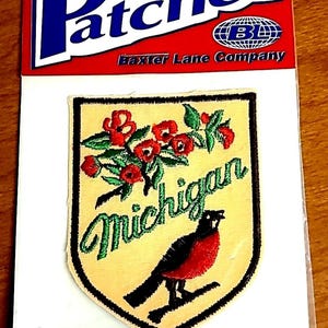 May include: Iron-on patch featuring a shield design with the word "Michigan" in green script. The patch includes a red bird, red flowers, and green leaves on a tan background. The top of the packaging reads "Patches" in blue and white.