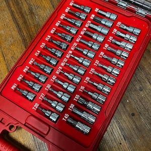May include: A red toolbox with a clear lid, filled with various metal screwdriver bits. The bits are organized in rows and labeled with sizes such as T20, T27, and H1/8. The toolbox is on a wooden surface.