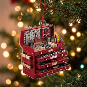 May include: A red tool chest ornament featuring a pegboard back with various miniature tools, multiple open drawers, and the text SARAH and 2026 printed on the front. The ornament hangs from a thin red string against a blurred holiday light background.