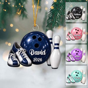 May include: A round bowling ball ornament featuring the name David and the year 2026 in white text, accompanied by a pair of blue and white bowling shoes and a white bowling pin, all suspended by a gold string against a blurred background.