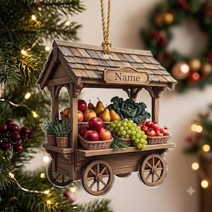 May include: A hanging wooden market stall ornament featuring a shingled roof with a sign reading Name. The stall displays miniature baskets filled with red apples, green grapes, pears, pomegranates, and leafy greens, supported by four wheels.
