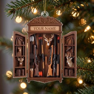 May include: A brown wooden cabinet-style Christmas ornament featuring open doors, displaying hunting gear including rifles, arrows, and deer heads. The top panel has the text YOUR NAME engraved in a light wood finish, with a gold hanging loop.