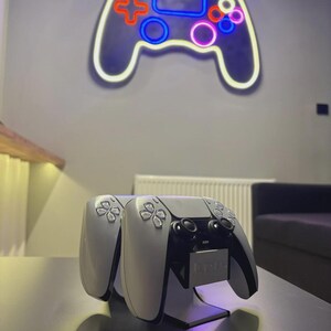 May include: A neon sign in the shape of a video game controller hangs on a gray wall. Below, two white and black game controllers rest on a black surface, with a stand. The neon sign has a white outline with red, blue, and pink accents.