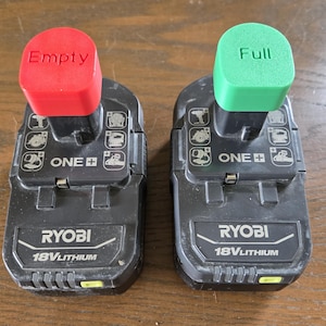 May include: Two black Ryobi 18V Lithium battery packs, one with a red cap labeled "Empty" and the other with a green cap labeled "Full". The packs have various tool icons and the "ONE+" logo.