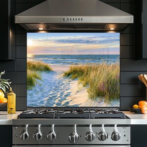 May include: A kitchen backsplash featuring a serene beach scene with a path leading to the ocean. The artwork depicts a sandy path, tall grasses, and a sunset over the water. The backsplash is installed behind a stainless steel range.