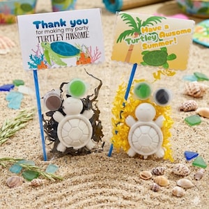 May include: Two clear bags with turtle-shaped craft kits on a sandy surface. Each kit includes a white turtle figurine, paint pots, and a sign that reads "Thank you for making my party TURTLEY AWSOME" and "Have a Turtley Awsome Summer!"