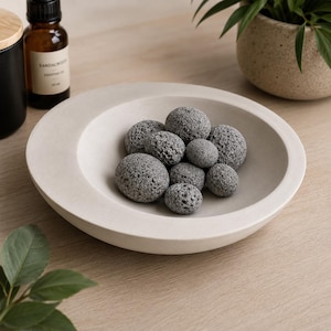 May include: A shallow, circular, off-white concrete bowl containing a collection of small, porous, dark gray volcanic stones. The bowl has a smooth, matte finish and a wide, flat rim, contrasting with the rough, textured surface of the stones.