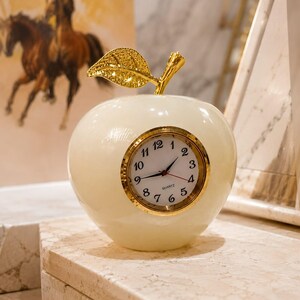 May include: An apple-shaped desk clock made of smooth, off-white stone with a polished finish. It features a gold-colored metal leaf at the top and a circular clock face with black Arabic numerals, black hour and minute hands, and a red second hand.