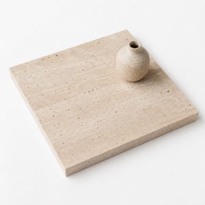 May include: A square beige travertine stone tile with a porous, natural surface texture. A small, round, light-colored ceramic vase with a narrow neck and a textured finish sits on the top right corner of the flat stone tile.