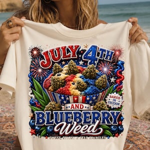 Può includere: T-shirt color crema con una grafica colorata che celebra il 4 luglio. Il design presenta un cupcake con glassa rossa, bianca e blu, circondato da fuochi d'artificio, foglie di cannabis e le parole "July 4th" e "Blueberry Weed."