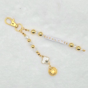 May include: Gold-colored keychain with a lobster clasp, featuring white and gold beads, and the word "OPALITE" spelled out in white letter beads. A charm with a cloud and lightning bolt hangs below a gold coin charm.