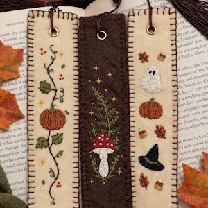 May include: Three rectangular felt bookmarks with dark brown tassels. The left one is cream with an embroidered pumpkin vine. The center one is dark brown with a red mushroom and stars. The right one is cream with a ghost, pumpkin, and hat.