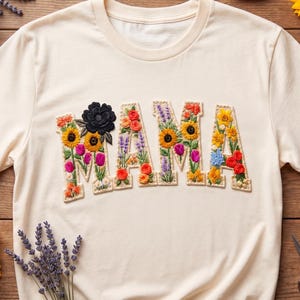 Puede incluir: Camiseta color crema con la palabra "MAMA" bordada con flores coloridas. La "M" presenta una flor negra. Girasoles, lavanda y suministros de costura rodean la camisa sobre una superficie de madera.