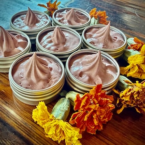 Puede incluir: Seis latas redondas de metal plateado rellenas de una crema lisa de color malva, cada una rematada con un remolino decorativo. Las latas están dispuestas en grupo y rodeadas de varias flores de caléndula amarillas y naranjas con tallos verdes.