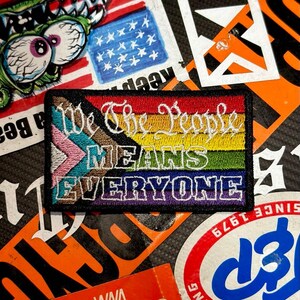 May include: A rectangular embroidered patch featuring the Progress Pride flag colors as a background. The white embroidered text reads We The People MEANS EVERYONE. The patch has a thick black border and is displayed against a background of stickers.
