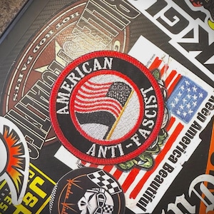May include: A round black patch with red and white trim. The patch features a stylized American flag and the words "AMERICAN ANTI-FASCIST" in white lettering. The patch is surrounded by other stickers.