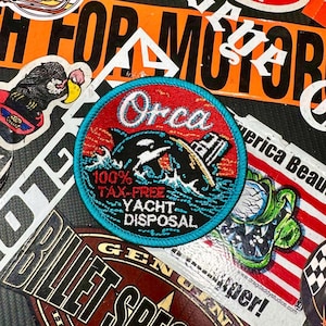 May include: A circular embroidered patch with a bright turquoise border. The center features a black orca whale jumping out of blue waves against a red background. It includes the white text Orca, 100% TAX-FREE, and YACHT DISPOSAL.