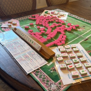 May include: A Mah Jongg game set featuring numerous pink and green tiles scattered across a green play mat. A wooden tile rack holds a row of tiles, while printed score cards and instruction sheets with text are arranged on the table surface.