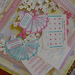 Puede incluir: Un juego de Mahjong con fichas rosas y blancas, cartas y un tablero de juego. El juego incluye fichas con diseños florales, tarjeteros rosas y hojas de puntuación. El tablero de juego tiene un patrón floral amarillo y rosa.