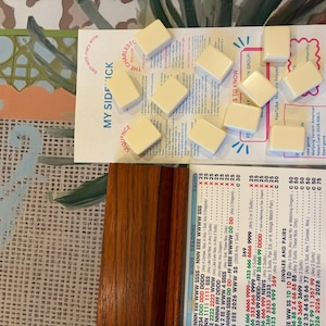Puede incluir: Un conjunto de diez fichas de mahjong de color blanco roto dispuestas sobre una tarjeta de instrucciones blanca titulada MY SIDESICK, junto a un soporte de madera y una hoja de referencia impresa con combinaciones y reglas de juego.