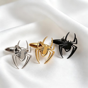 Puede incluir: Tres gemelos con forma de araña en plata, oro y negro. Los gemelos se exhiben sobre una superficie blanca y sedosa. Cada gemelo presenta un diseño detallado de araña, perfecto para añadir un toque único.